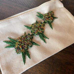 May include: A cream-colored zippered pouch with three embroidered cannabis bud designs. The buds are detailed with green, gold, and purple thread, and are surrounded by green embroidered leaves. The pouch is made of a textured fabric.
