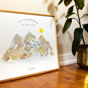 May include: A framed print with a mountain range design made from vintage map pieces. The artwork includes the text "LIFE IS AN ADVENTURE BEST TRAVELLED TOGETHER" and a yellow sun. The frame is made of light-coloured wood.
