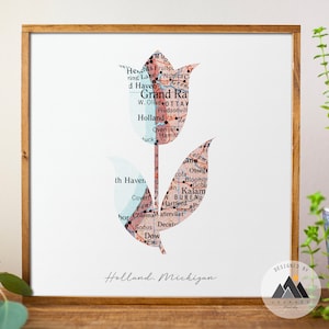 Puede incluir: Una impresión enmarcada de una flor de tulipán hecha a partir de un mapa de Holland, Michigan. El tulipán es rosa y azul y el texto "Holland, Michigan" está impreso debajo de la flor. La impresión está enmarcada en un marco de madera claro.