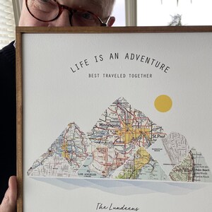 May include: A framed print with a white background and a map design. The map is made up of three mountains, each with a different city name. The text "Life is an adventure, best traveled together" is at the top of the print. The text "The Lundeens, Est. 1992" is at the bottom of the print.