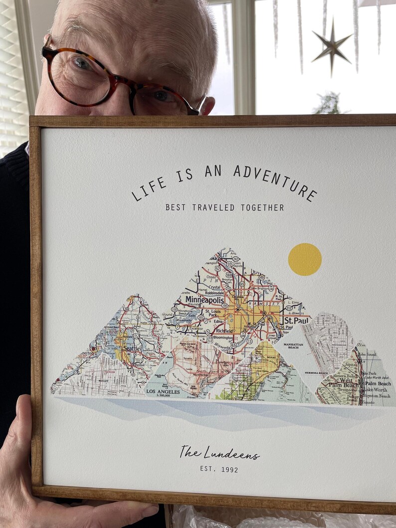 May include: A framed print with a white background and a map design. The map is made up of three mountains, each with a different city name. The text "Life is an adventure, best traveled together" is at the top of the print. The text "The Lundeens, Est. 1992" is at the bottom of the print.