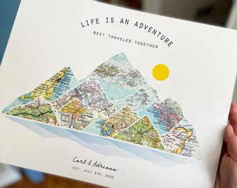 May include: A white print with a mountain range made of vintage maps. The mountains are surrounded by a blue watercolor effect. The text "LIFE IS AN ADVENTURE" is at the top of the print. The text "BEST TRAVELED TOGETHER" is below the first line of text. The text "Carl & Adriana EST. JULY 4TH, 2020" is at the bottom of the print.