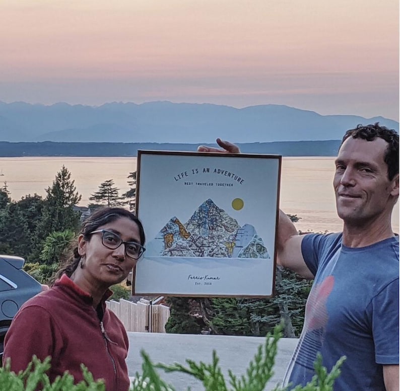 May include: A couple poses in front of a mountain range with a framed print that reads "Life is an adventure, best traveled together." The print features a stylized mountain range with a sun and the text "Farris & Lamar Est. 2018."