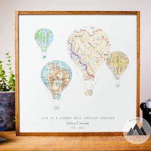 A framed wooden panel rests on a shelf. On the print are hot air balloons made up of different maps. Below the balloons reads the customizable words Life is a journey best traveled together. Custom text below includes 2 names and a date.