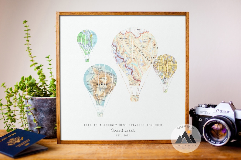 A framed wooden panel rests on a shelf. On the print are hot air balloons made up of different maps. Below the balloons reads the customizable words Life is a journey best traveled together. Custom text below includes 2 names and a date.
