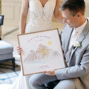 May include: A groom holds a framed print with a mountain range and the text "You are my home and adventure all at once." The print is a wedding gift for the bride.