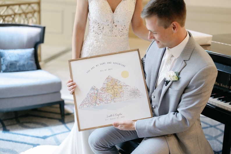 May include: A groom holds a framed print with a mountain range and the text "You are my home and adventure all at once." The print is a wedding gift for the bride.