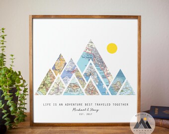 May include: A framed print with a mountain range made of vintage maps. The mountains are surrounded by a light blue background with a yellow sun. The text "Life is an adventure best traveled together" is at the bottom of the print, along with the names "Michael & Stacy" and the date "EST. 2017".