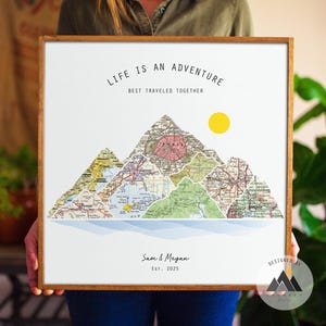 Könnte beinhalten: Gerahmter Druck mit Bergformen aus alten Kartenstücken. Der Text lautet "LIFE IS AN ADVENTURE BEST TRAVELED TOGETHER" und "Sam & Megan Est. 2025". Eine gelbe Sonne ist abgebildet. Der Rahmen ist braun.