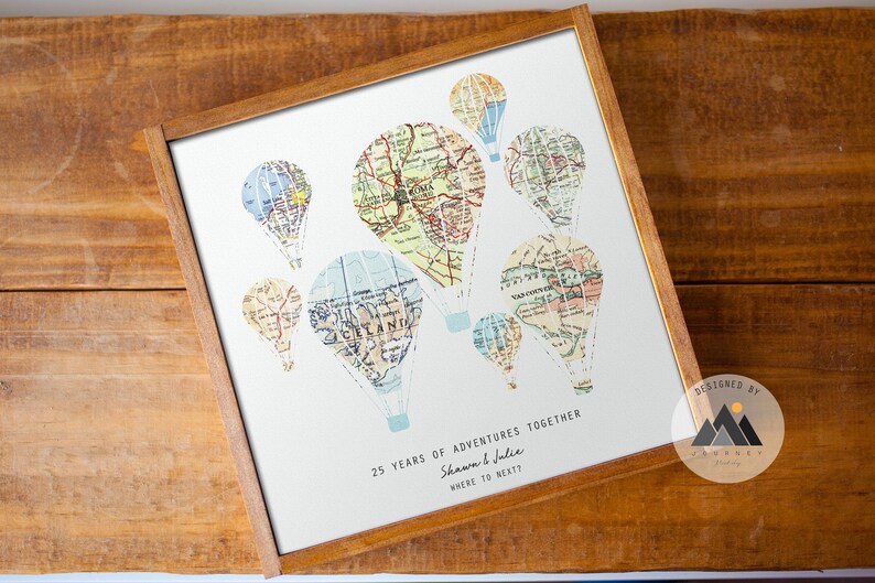 May include: A framed print with a white background featuring a design of hot air balloons made from vintage maps. The balloons are in various colors and sizes. The text below the balloons reads "25 years of adventures together Shannon & Julie Where to next?" and "Designed by Journey's Studio".