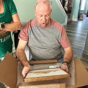 May include: A person is opening a brown cardboard box to reveal a framed print with a colorful image. The print is in a wooden frame with a gold trim.