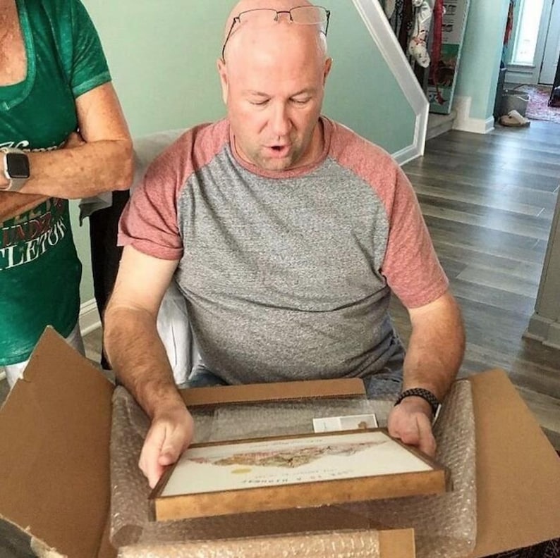 May include: A person is opening a brown cardboard box to reveal a framed print with a colorful image. The print is in a wooden frame with a gold trim.