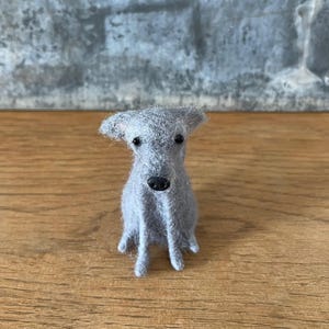 May include: A handmade, gray felt dog figurine with a black nose and small, black eyes. The dog is sitting upright on a wooden surface. The background is a gray, textured wall.