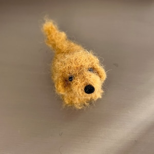 Puede incluir: Un pequeño perro de fieltro de aguja marrón con ojos negros y nariz negra. El perro está acostado con la cabeza girada hacia un lado.