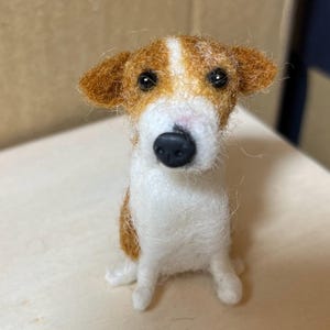 Op de afbeelding: Handgemaakte vilten hondenfiguur in zittende positie. De hond heeft een wit lichaam met bruine en bruine aftekeningen op zijn hoofd en oren. Hij heeft een zwarte neus en donkere ogen, wat hem een realistisch uiterlijk geeft. Een charmant decoratief item.