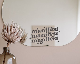 May include: A teardrop-shaped mirror with a white background and the word "manifest" repeated three times in a stylized font. The mirror is hanging on a light pink wall above a wooden table with a vase of dried flowers, a white ceramic sculpture, and a stack of books.