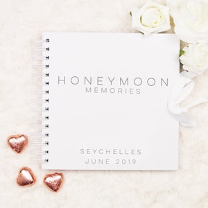 May include: White spiral bound notebook with the text "Honeymoon Memories" and "Seychelles June 2019" on the cover. The notebook is surrounded by white roses and small gold heart-shaped chocolates.