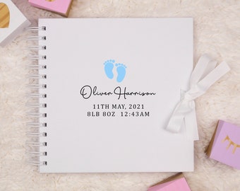 May include: A white baby memory book with a blue footprint design. The book has a white ribbon and the text "Oliver Harrison, 11th May, 2021, 8lb 8oz 12:43AM" printed on the front cover.