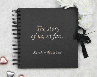 May include: Black scrapbook with a spiral binding and a black ribbon. The cover has the text "The story of us, so far..." and "Sarah + Matthew" in a rose gold font.
