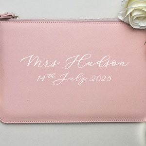 May include: A pale pink zippered pouch with a white inscription that reads "Mrs. Hudson 14th July 2025".