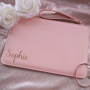 May include: A light pink clutch with a gold zipper and a gold ring handle. The clutch has the name "Sophie" written in gold script on the front.