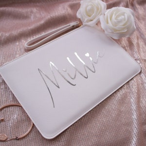 May include: White clutch bag with a silver metallic inscription that reads "Millie" with a small heart.