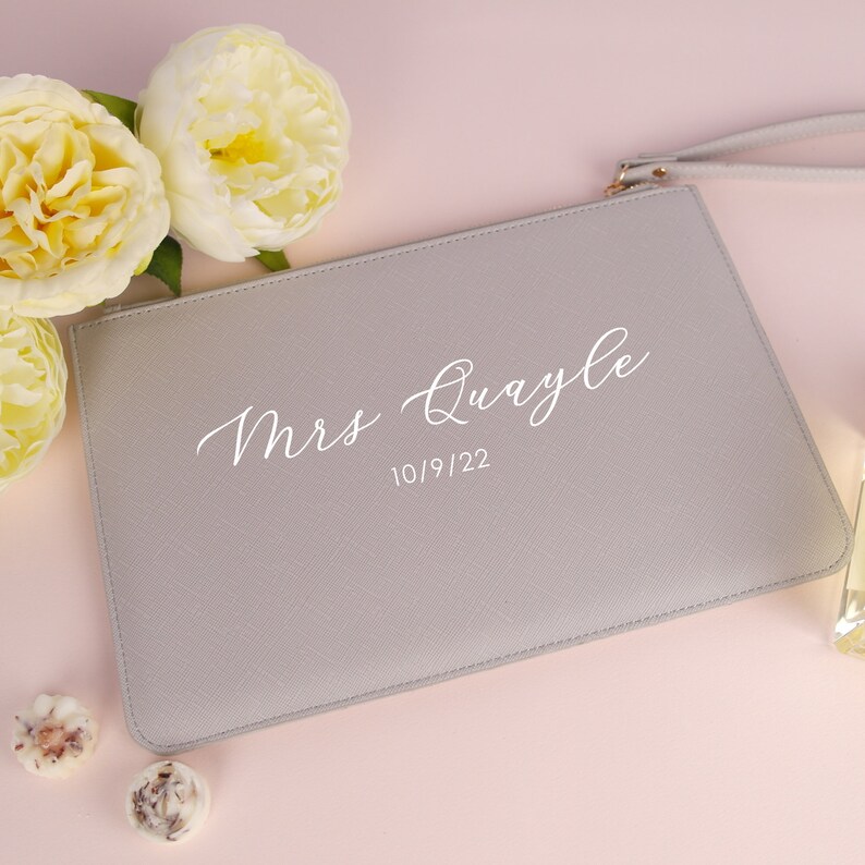 May include: A grey clutch bag with a white zipper and a white strap. The bag has the text "Mrs. Quayle" and the date "10/9/22" printed on it.