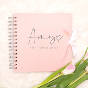 May include: Pink scrapbook with a white ribbon and the text "Amy's Hen Weekend" on the cover. The scrapbook is lying on a white fluffy surface with a bouquet of white tulips.