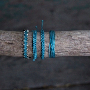 Puede incluir: Cinco pulseras de color azul turquesa sobre una superficie de madera. Las pulseras están hechas de diferentes materiales, incluyendo cordón, cuentas y tejido tejido.
