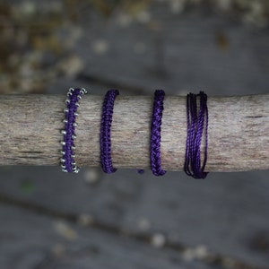 Puede incluir: Cinco pulseras moradas, incluida una con cuentas plateadas, se muestran en un trozo de madera a la deriva.
