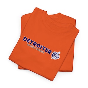 May include: Two orange t-shirts are folded and stacked. The top shirt displays the text "DETROITER MOBILE HOMES" in blue, along with a blue and white logo. The shirts are made of a soft material.