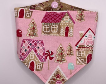 May include: A pink dog bandana with a gingerbread house pattern. The bandana features gingerbread houses, trees, and candy canes in shades of pink, brown, and red. The reverse side is brown with a gingerbread pattern and fastens with a pink snap.