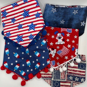 May include: Five different fabric bandanas with a patriotic theme. The bandanas are red, white, and blue with stars, stripes, and a dog wearing sunglasses. The bandanas have pom-poms around the edges.