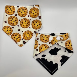 May include: Two reversible bandana bibs for babies or toddlers. One side features a white background with a brown and yellow chocolate chip cookie pattern. The other side features a white and black cow print pattern. The bibs have two white snap buttons.