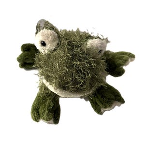May include: A plush toy in the shape of a green alligator. The toy has a shaggy, textured body, white eyes, and dark green feet. The alligator is lying flat on a white background.