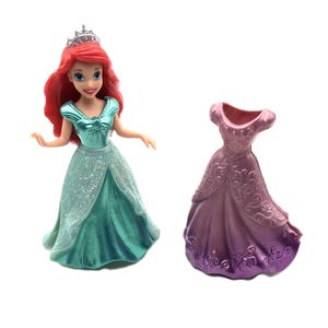 May include: A plastic doll of Ariel from Disney's The Little Mermaid wearing a teal dress and a silver crown. The doll is holding a separate purple dress with a floral pattern.
