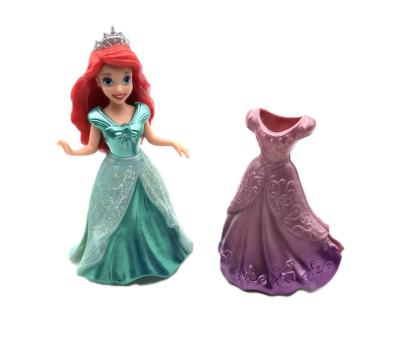 May include: A plastic doll of Ariel from Disney's The Little Mermaid wearing a teal dress and a silver crown. The doll is holding a separate purple dress with a floral pattern.