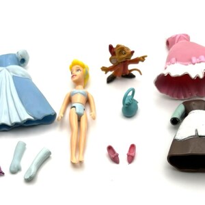 Op de afbeelding: Een verzameling speelgoed met het thema Assepoester, waaronder een pop, jurken, schoenen en accessoires. De jurken zijn blauw, roze en bruin. Een kleine muisfiguur en een miniatuur portemonnee zijn ook inbegrepen. De pop heeft geel haar en draagt een blauwe bikini.