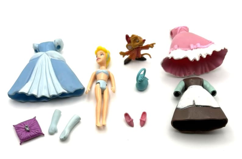 Op de afbeelding: Een verzameling speelgoed met het thema Assepoester, waaronder een pop, jurken, schoenen en accessoires. De jurken zijn blauw, roze en bruin. Een kleine muisfiguur en een miniatuur portemonnee zijn ook inbegrepen. De pop heeft geel haar en draagt een blauwe bikini.