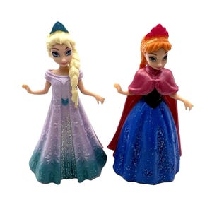 Pu&ograve; includere: Due statuette di personaggi animati. Una ha i capelli biondi in una treccia e indossa un vestito lilla chiaro con un effetto ombretto blu-verde. L'altra ha i capelli rossi e indossa un vestito blu con un mantello rosso.