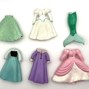 Op de afbeelding: Zes miniatuur prinsessenjurkfiguren in verschillende kleuren. Inclusief een groene rok, een witte jurk met blauwe accenten, een groene zeemeerminstaart, een blauwgroene jurk met een zwart lijfje, een paarse jurk en een roze jurk.