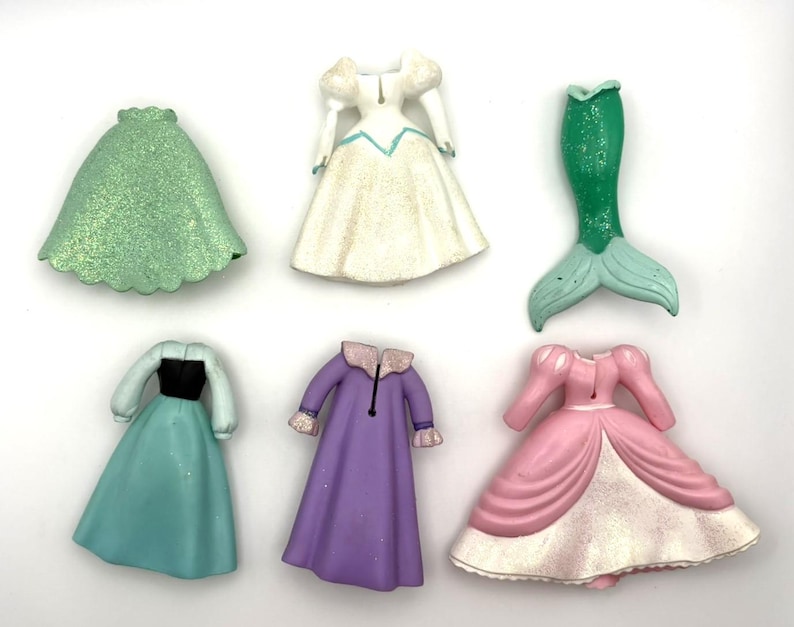 Op de afbeelding: Zes miniatuur prinsessenjurkfiguren in verschillende kleuren. Inclusief een groene rok, een witte jurk met blauwe accenten, een groene zeemeerminstaart, een blauwgroene jurk met een zwart lijfje, een paarse jurk en een roze jurk.