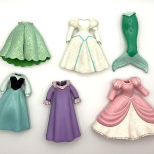Op de afbeelding: Een verzameling miniatuur prinsessenjurken en een zeemeerminstaart in verschillende kleuren. De jurken omvatten een groene rok, een witte jurk met blauwe accenten, een turquoise jurk met een zwart lijfje, een paarse jurk en een roze jurk met witte rand.