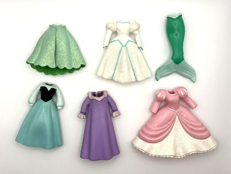 Op de afbeelding: Een verzameling miniatuur prinsessenjurken en een zeemeerminstaart in verschillende kleuren. De jurken omvatten een groene rok, een witte jurk met blauwe accenten, een turquoise jurk met een zwart lijfje, een paarse jurk en een roze jurk met witte rand.