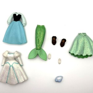 May include: A selection of doll clothing and accessories, including a blue dress with black bodice, a white dress with blue accents, a green mermaid tail, brown shoes, and a light blue ring. The items are arranged on a white surface.
