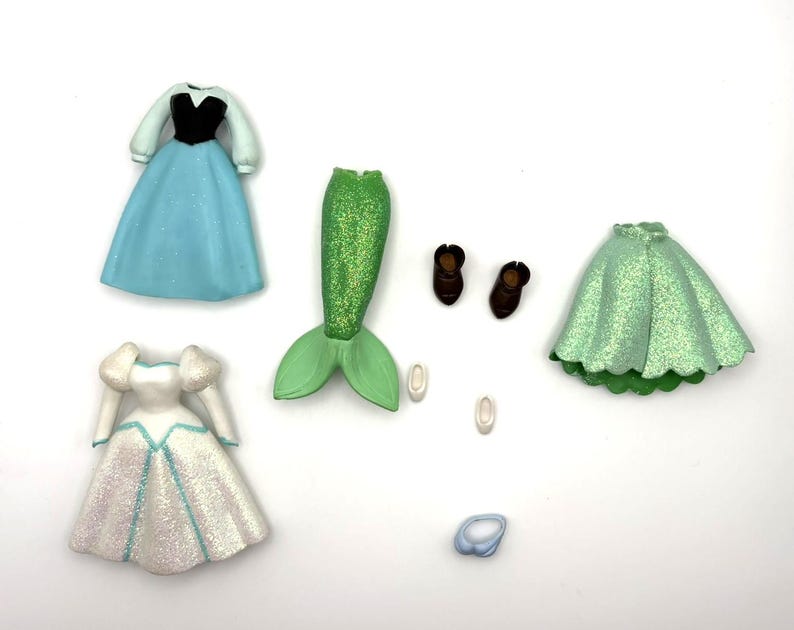 May include: A selection of doll clothing and accessories, including a blue dress with black bodice, a white dress with blue accents, a green mermaid tail, brown shoes, and a light blue ring. The items are arranged on a white surface.
