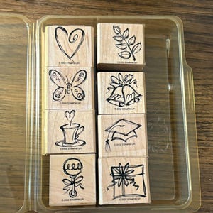 May include: A set of eight square wooden rubber stamps arranged in a clear plastic storage case. Each stamp features a black line art illustration including a heart, a leaf sprig, a butterfly, two bells, a coffee cup, a graduation cap, a baby rattle, and a gift box. Each stamp is marked with the text 2002 STAMPIN'UP!