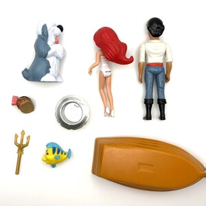 Op de afbeelding: Een verzameling Disney's De Kleine Zeemeermin beeldjes en accessoires. Inclusief Ariel, Prins Eric, Botje, een boot, een hond, een drietand en andere kleine items. De figuren zijn gemaakt van plastic en zijn felgekleurd.