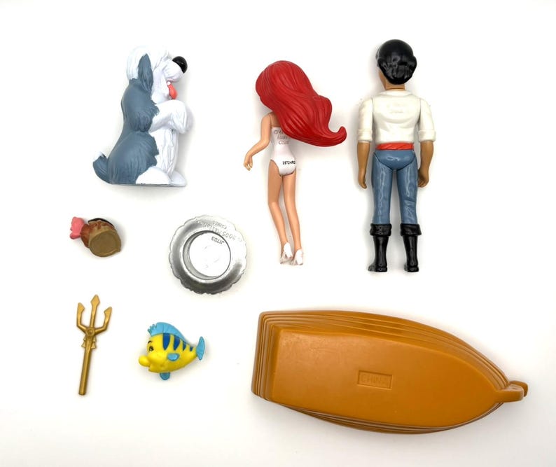 Op de afbeelding: Een verzameling Disney's De Kleine Zeemeermin beeldjes en accessoires. Inclusief Ariel, Prins Eric, Botje, een boot, een hond, een drietand en andere kleine items. De figuren zijn gemaakt van plastic en zijn felgekleurd.