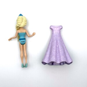 Pu&ograve; includere: Una bambola con i capelli biondi e un vestito blu &egrave; accanto a un vestito lilla chiaro. I capelli della bambola sono acconciati a treccia e l'abito ha un motivo floreale. La bambola indossa scarpe blu.